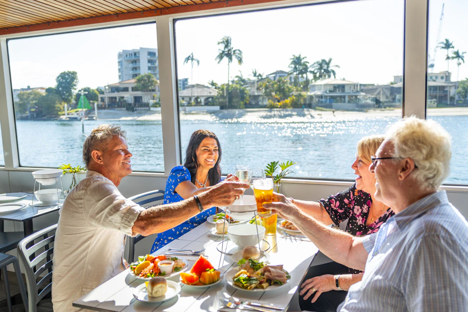 Three people dining on a boat, clinking drinks over a table with plates of food. Waterfront homes and palm trees are visible outside.
