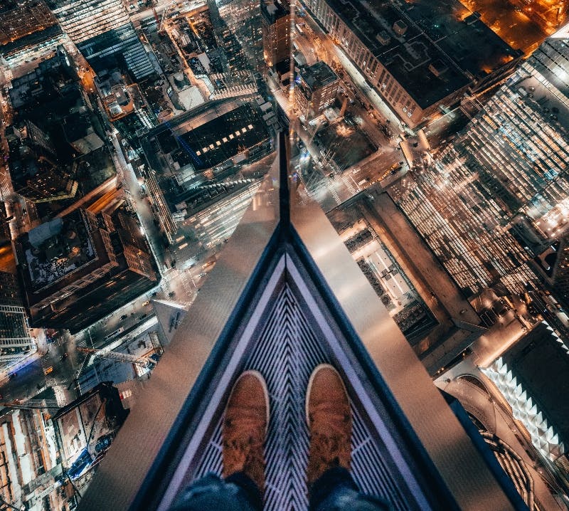 높은 건물 가장자리에 갈색 부츠를 신고 서서 밤에는 밝게 빛나는 도시 풍경이 내려다보입니다.