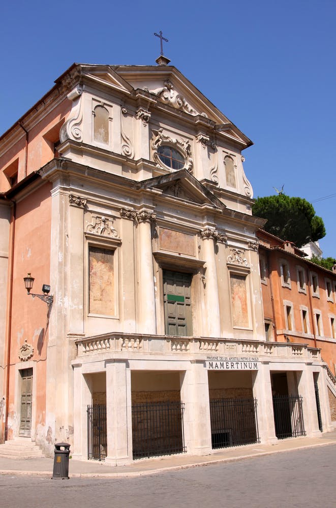 Historic building with ornate facade and green door, labeled "Mamertinum," next to a rustic orange building and a tree in the background.