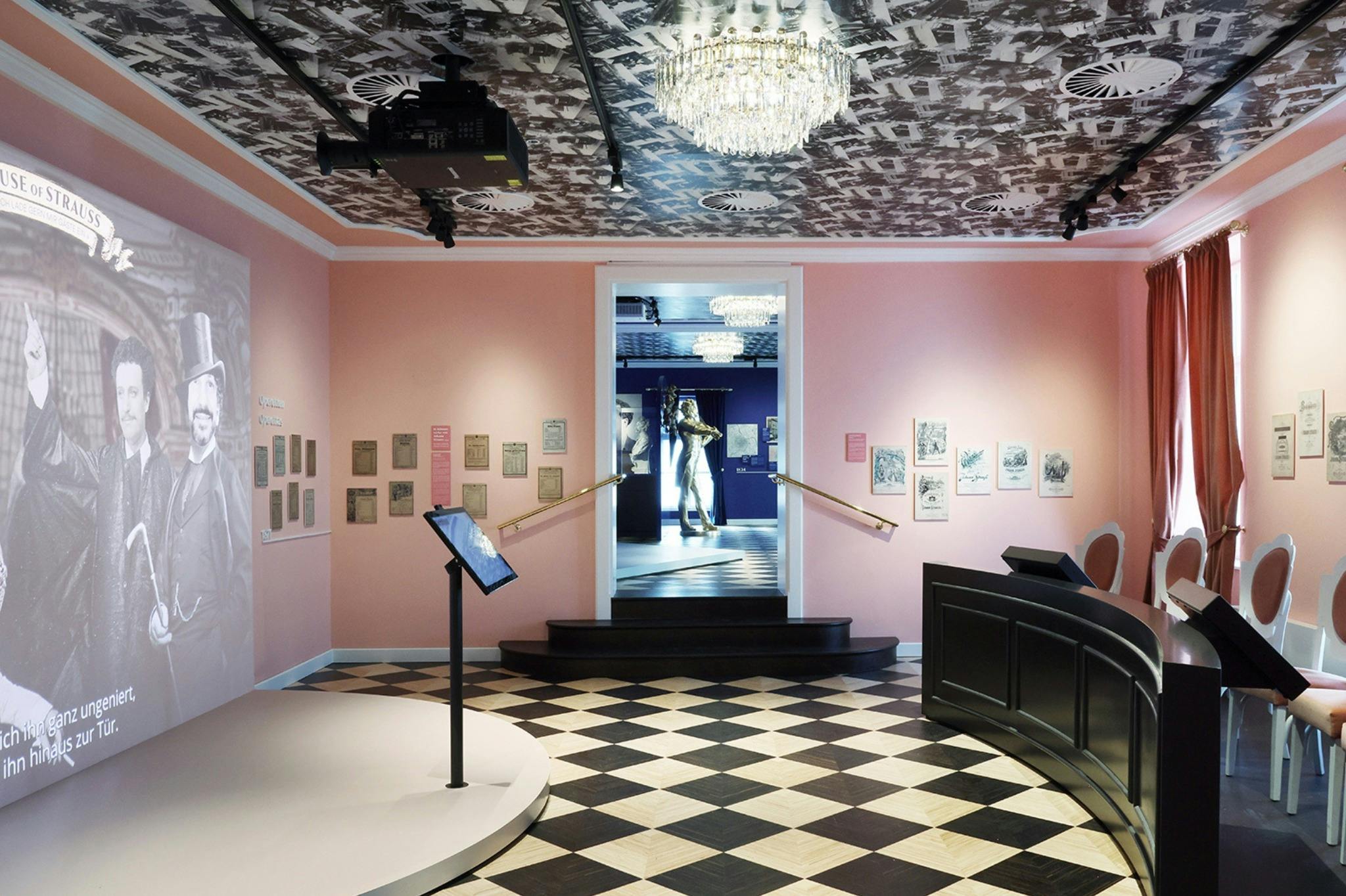 Eine Museumsausstellung mit rosafarbenen Wänden, einem karierten Boden, einem Kronleuchter, gerahmten Kunstwerken und einer Schaufensterpuppe im Hintergrund.