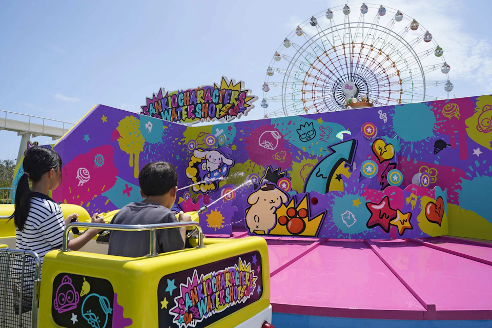 A child in a yellow ride vehicle at a colorful water-themed amusement park attraction, with a Ferris wheel in the background.