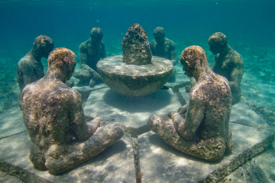 Underwater statues of people sitting in a circle around a central round structure.