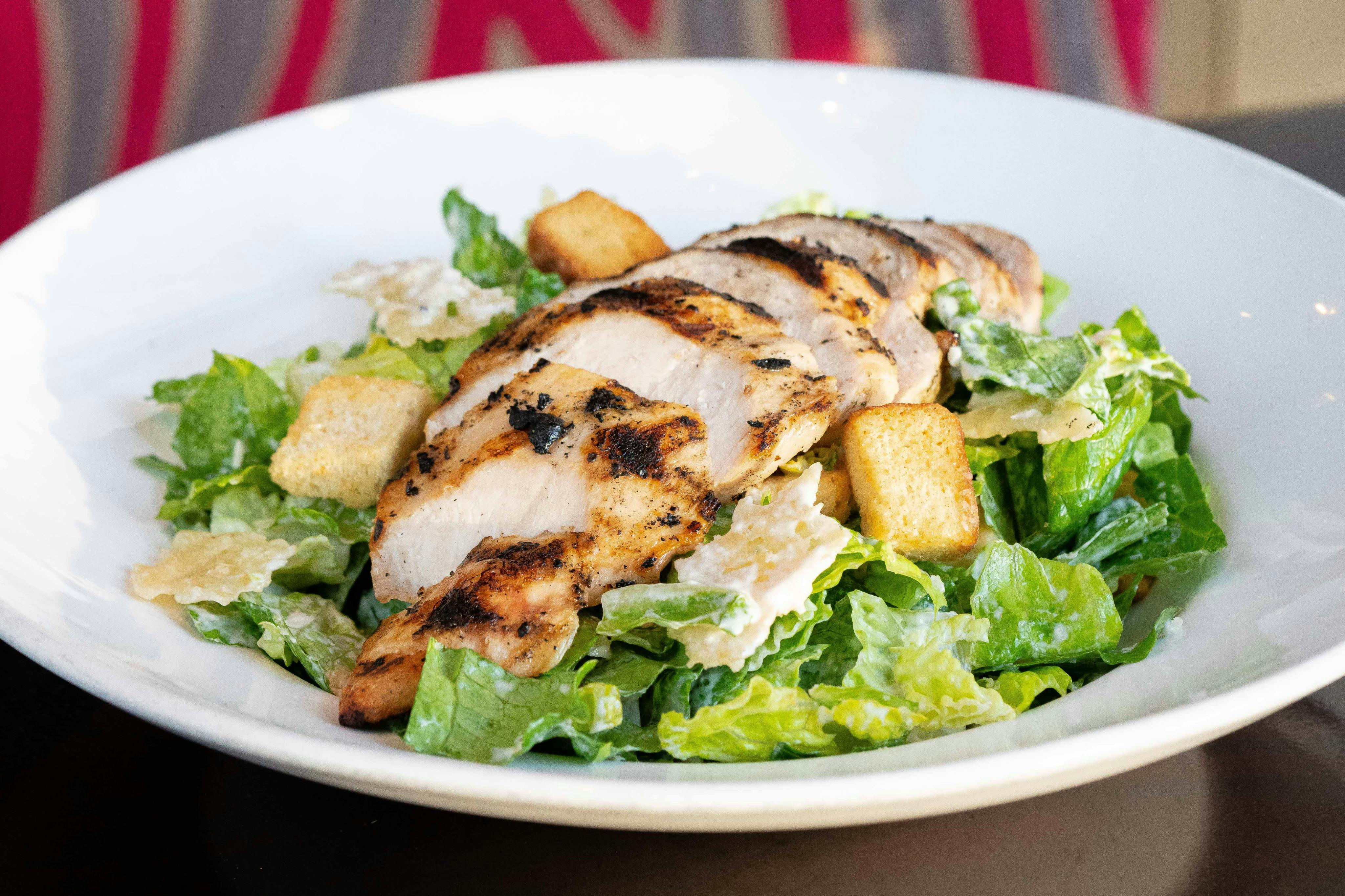 Grilled chicken slices on a bed of romaine lettuce with croutons and shaved Parmesan in a white bowl.