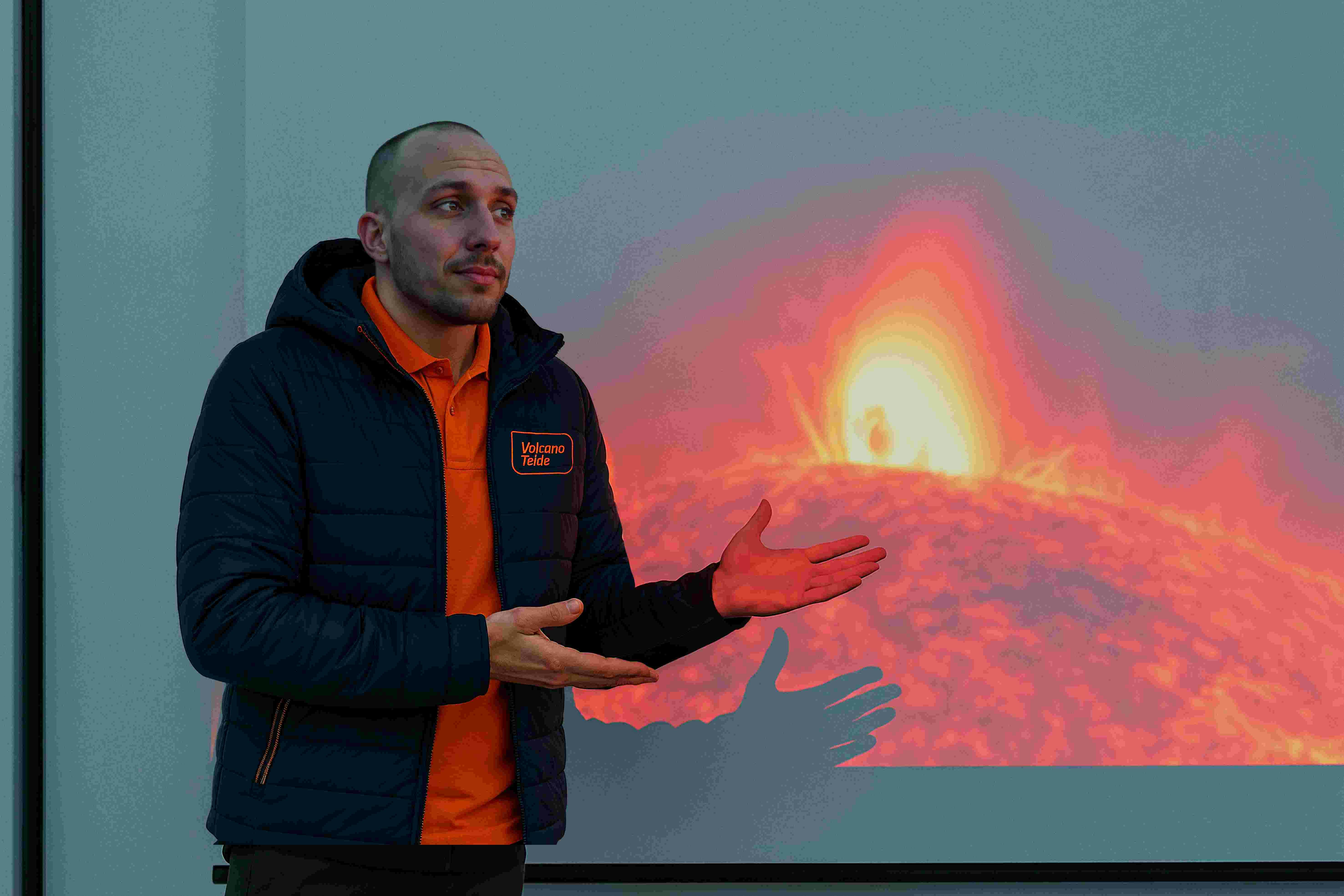 Man in a blue jacket and orange shirt speaks, gesturing toward a projected image of a volcanic eruption.