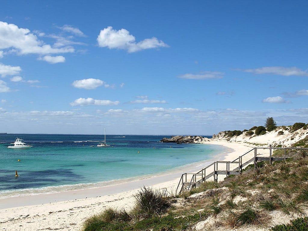 A sandy beach with clear blue water, several boats, a wooden staircase leading to the shore, and a partly cloudy sky overhead.