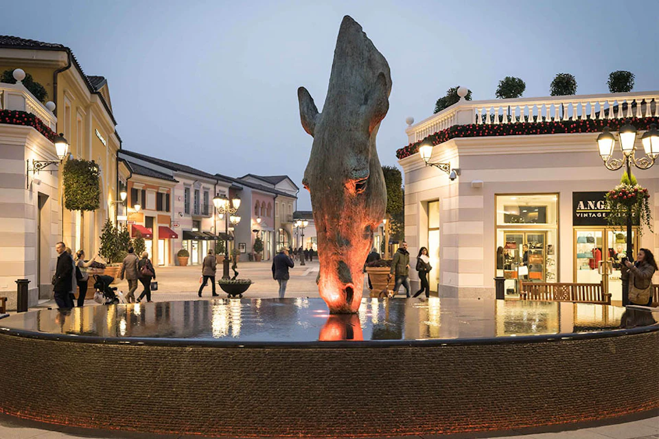 People walk past a large horse head sculpture in a plaza, surrounded by shops and lit by street lamps.
