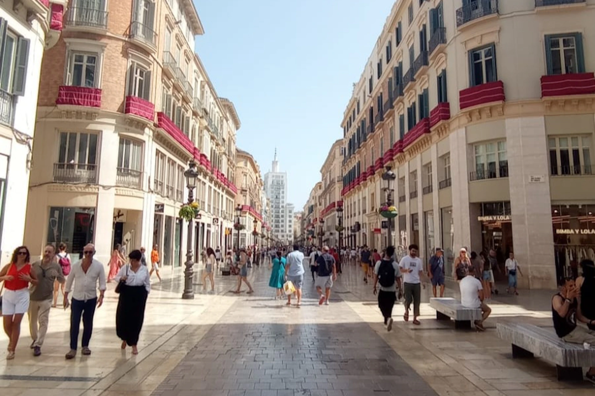 Pedestrian street lined with tall buildings, bustling with people. Red awnings adorn the windows, and the sky is clear.