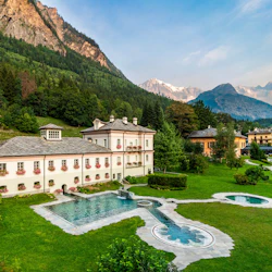 A luxurious villa with a pool, surrounded by green lawns and trees, set against a backdrop of mountains and a blue sky.