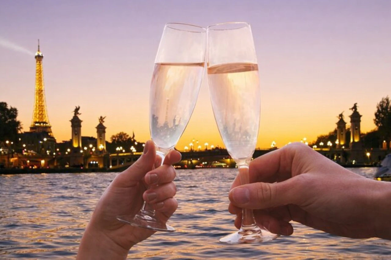 Two people clinking champagne glasses over a water scene at sunset, with a bridge and sculptures in the background.