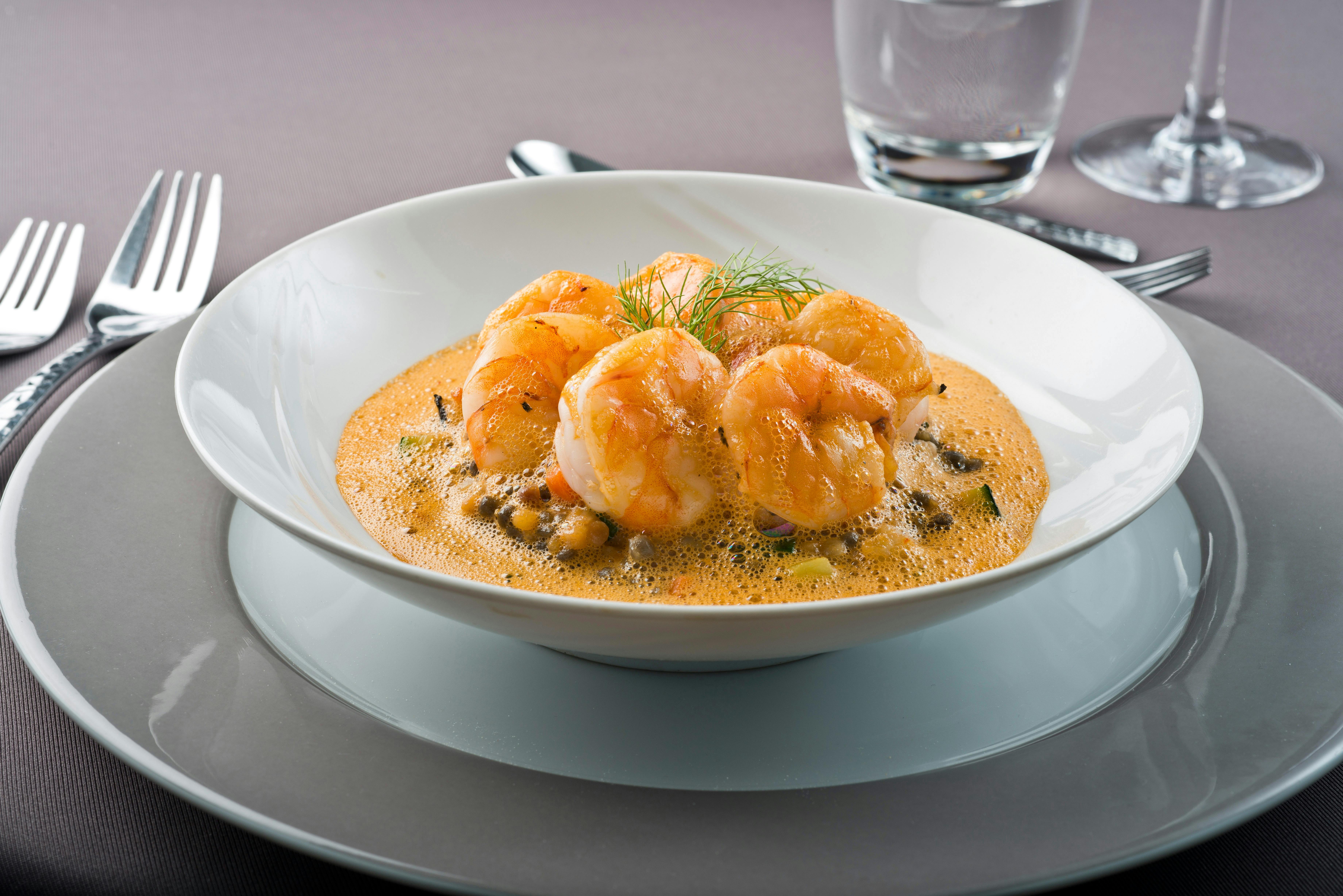 虾放在奶油泡沫酱中，用白色碗装饰香草，放在灰色盘子上。 背景中有一杯水。
