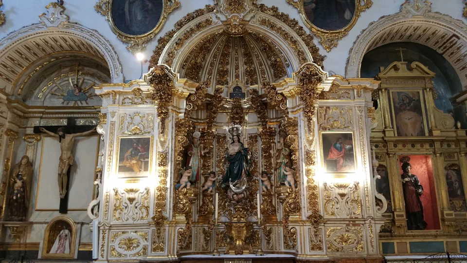 Ornate altar with a central statue of Mary, angels, two side paintings, and elaborate gold detailing in a church.