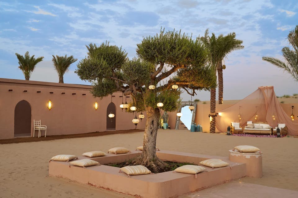 A courtyard with a tree adorned with lanterns, surrounded by pillows on a sandy ground, with palm trees and a building in the background.