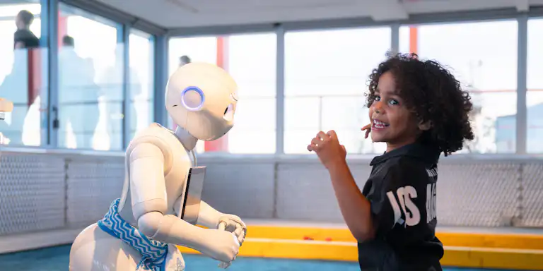 Et lille barn interagerer med en humanoid robot i et indendørs rum med store vinduer og flere mennesker i baggrunden.