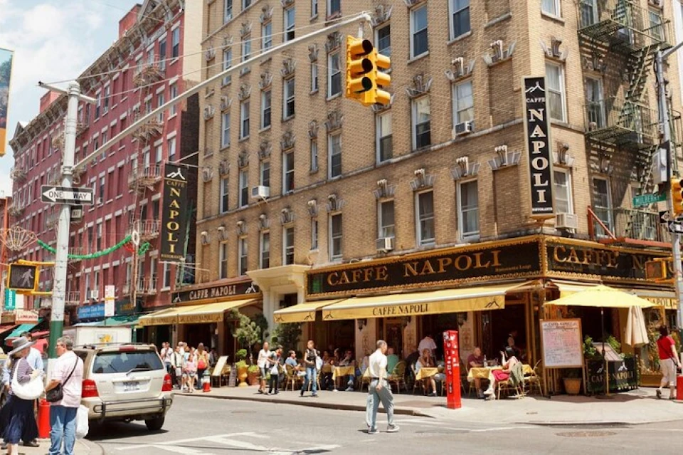 Street-side café with yellow awnings, outdoor seating, and people dining. Brown brick building and traffic light overhead.