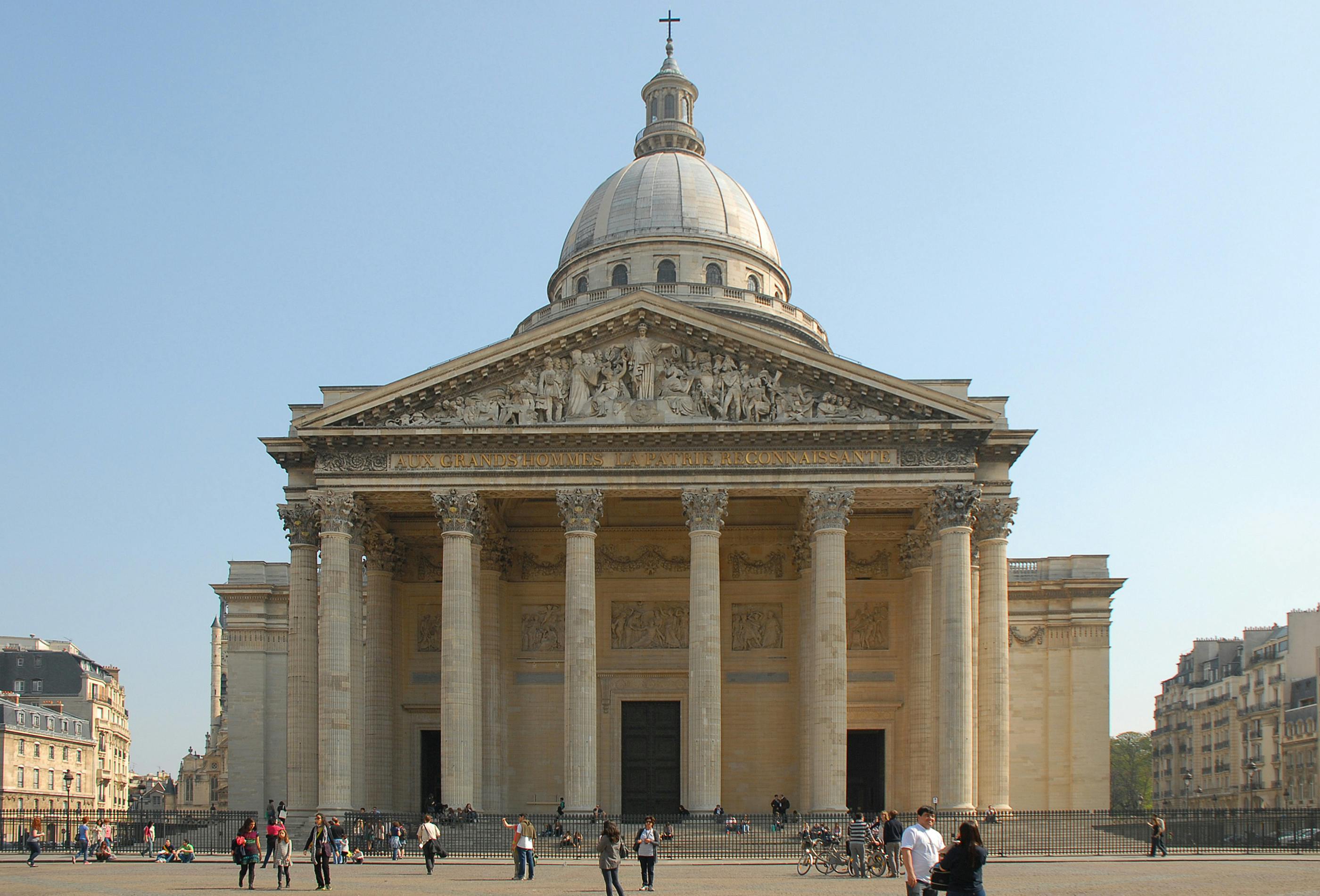 Large neoclassical building with a dome, fronted by six columns and detailed carvings. People and bikes are visible in the plaza.