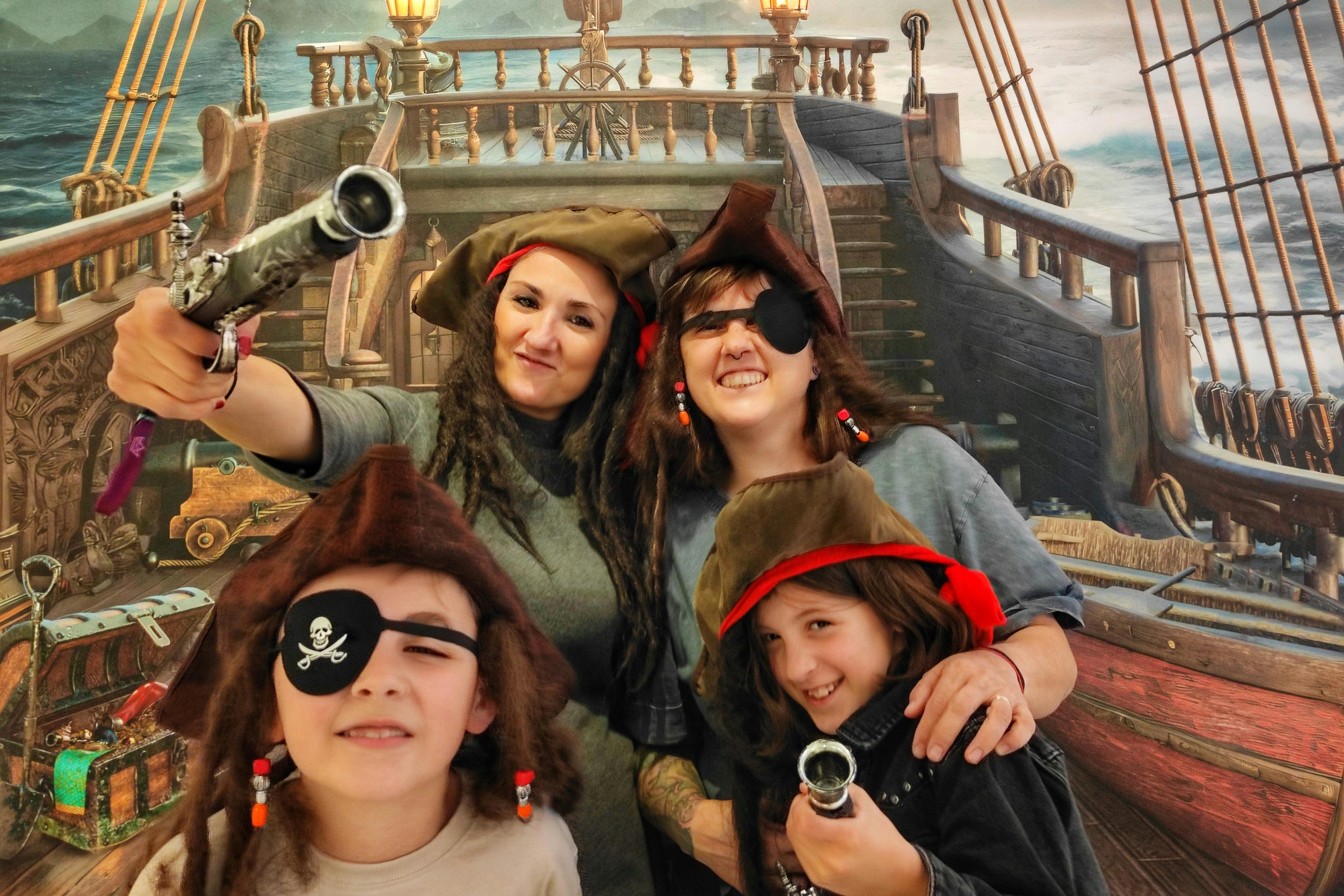 Τέσσερις άνθρωποι ντυμένοι πειρατές, φορώντας καπέλα, καλύμματα ματιών και κρατώντας όπλα-παιχνίδια, ποζάρουν μπροστά από ένα φόντο πλοίου.