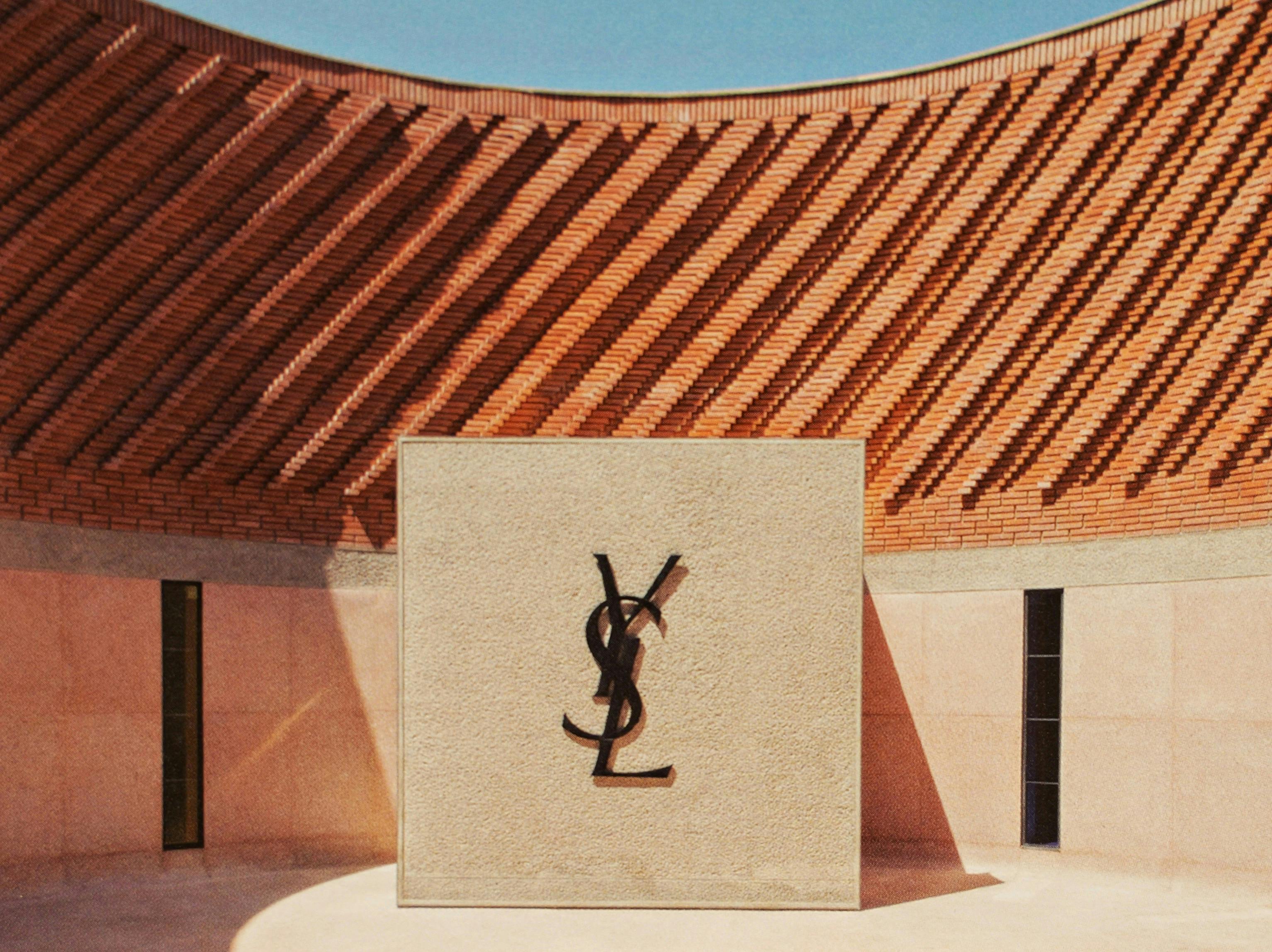 Un mur carré beige, orné d'un monogramme noir entrelacé, se dresse dans une cour entourée de motifs de briques rouges et sous un ciel bleu.