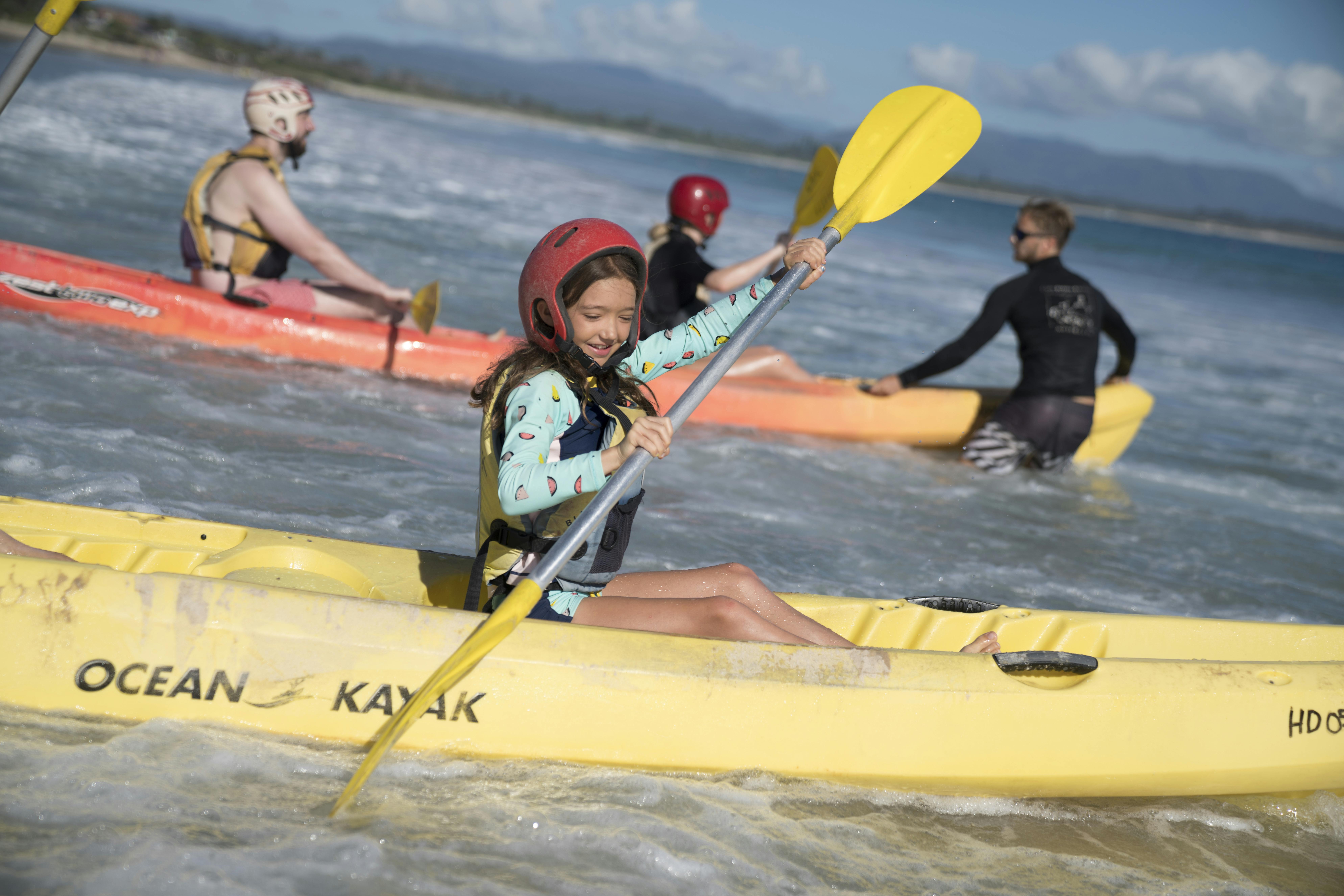 A child with a red helmet paddles a yellow kayak in the ocean, accompanied by two adults in orange kayaks.