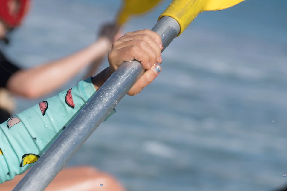 A child with a red helmet paddles a yellow kayak in the ocean, accompanied by two adults in orange kayaks.