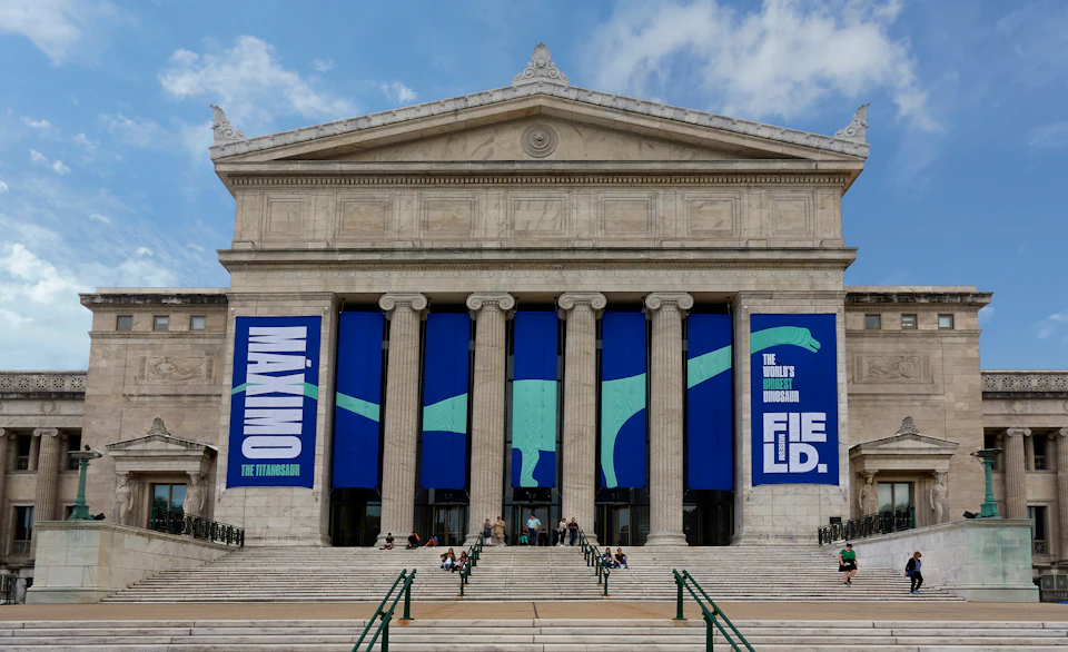 Facade of a neoclassical museum with large blue banners advertising "MAXIMO THE TITANOSAUR" and "THE WORLD'S BIGGEST DINOSAUR" exhibit.