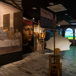 Museiutställning med Leonardo da Vincis uppfinningar, med modeller, stora målningar och en bakgrund av ett ljust landskap.