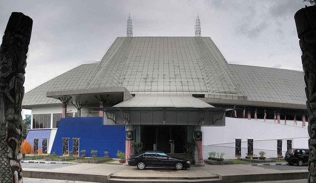 A large, modern building with a steeply sloped roof, flag decorations, and two parked cars in front of an entrance.