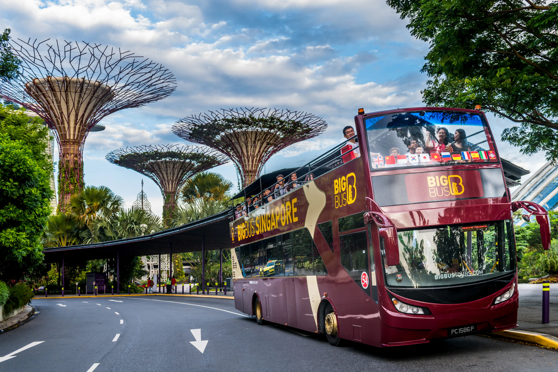 A red double-decker tour bus with "Hippo Singapore" signage drives by the Marina Bay Sands hotel under a partly cloudy sky.
