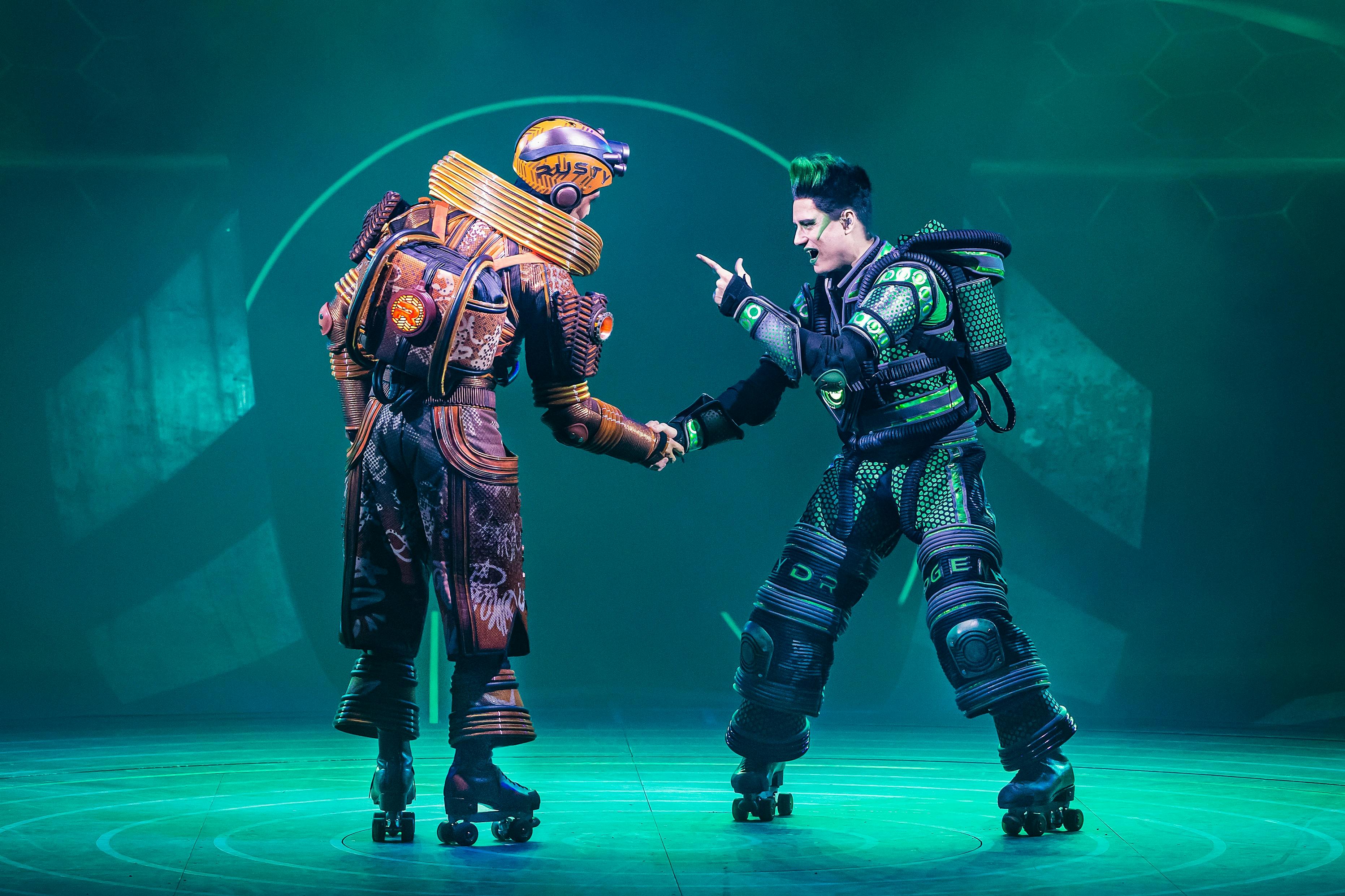 两名穿着溜冰鞋的表演者穿着色彩缤纷的未来服装和头盔，在绿灯照明的舞台上握手。