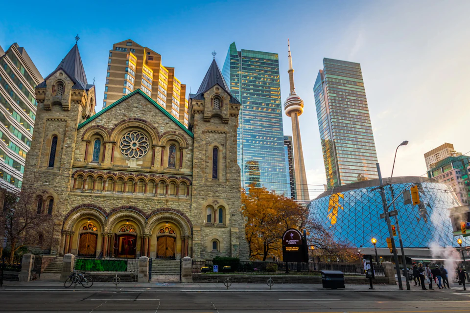A historic stone church with arched doors and windows stands in front of modern skyscrapers, including a tower with a CN Tower.
