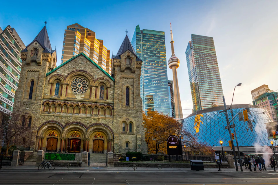 A historic stone church with arched doors and windows stands in front of modern skyscrapers, including a tower with a CN Tower.