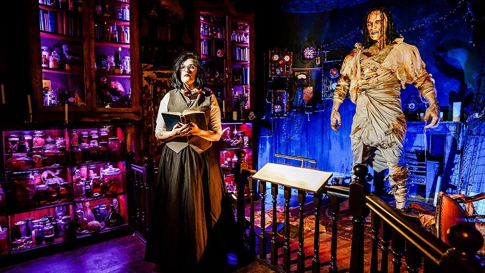 Victorian dressed woman holding book in spooky lab. Lifesize Frankenstein's monster in the background