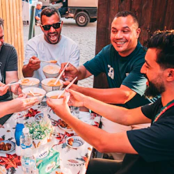 Four men sit at a table outdoors, smiling and sharing bowls of soup, with various foods and beverages on the table.