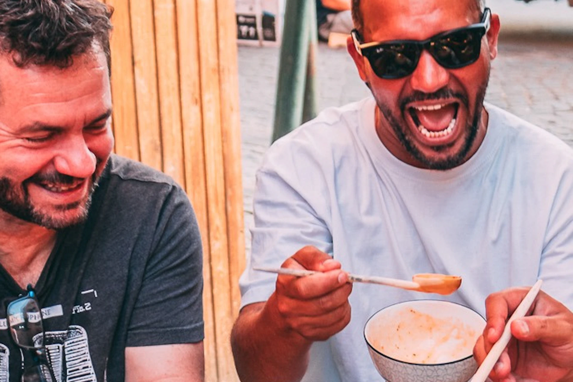 Four men sit at a table outdoors, smiling and sharing bowls of soup, with various foods and beverages on the table.