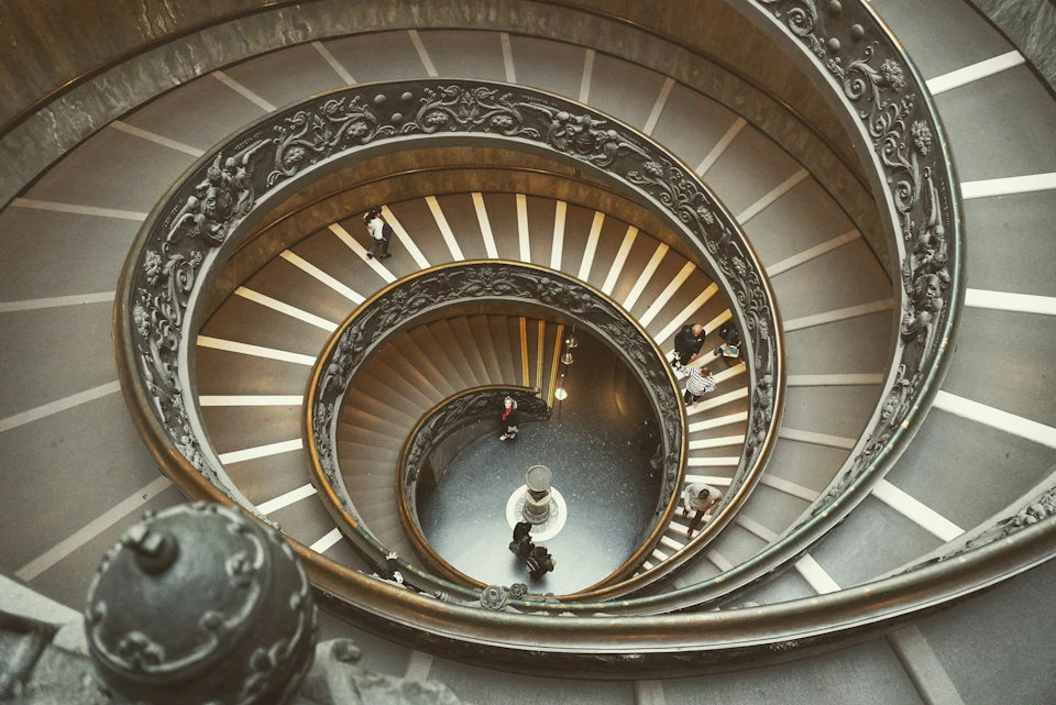 Spiral staircase with ornate railing viewed from above. People descending and ascending. Center features a small statue.