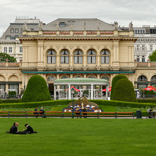 En park med människor som sitter på gräs och bänkar, framför en stor, utsmyckad byggnad med träd och klippta häckar.