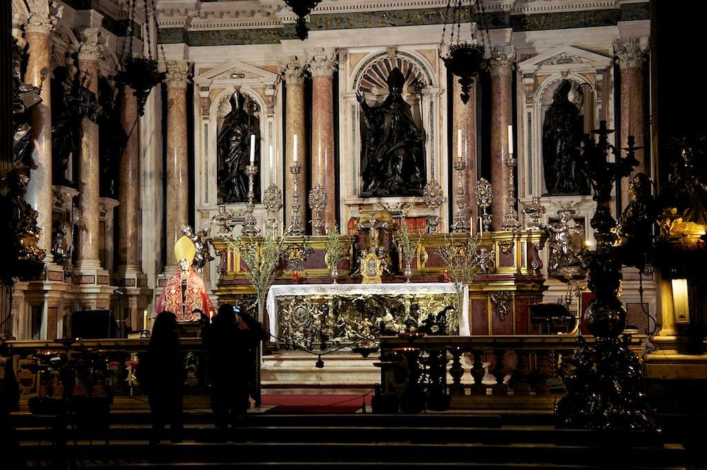 Μεγάλος βωμός σε έναν καθεδρικό ναό με περίτεχνα διακοσμητικά, αγάλματα και μανουάλια. Σιλουέτες δύο ατόμων σε πρώτο πλάνο.