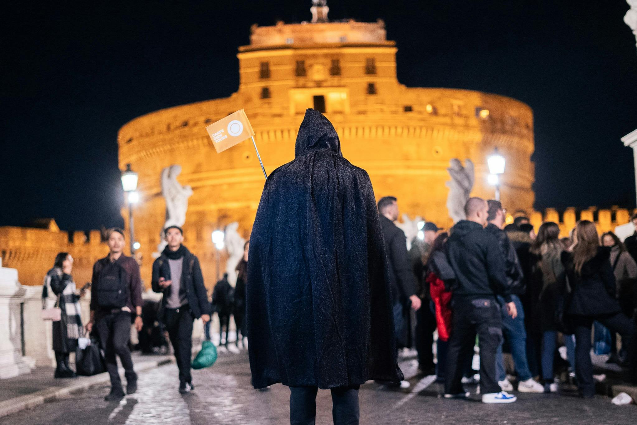 A person in a dark cloak holding a small flag stands on a cobblestone street at night, with a lit historic building in the background.