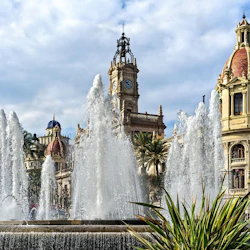 Fontän med höga vattenstrålar framför utsmyckade historiska byggnader under en molnig himmel.