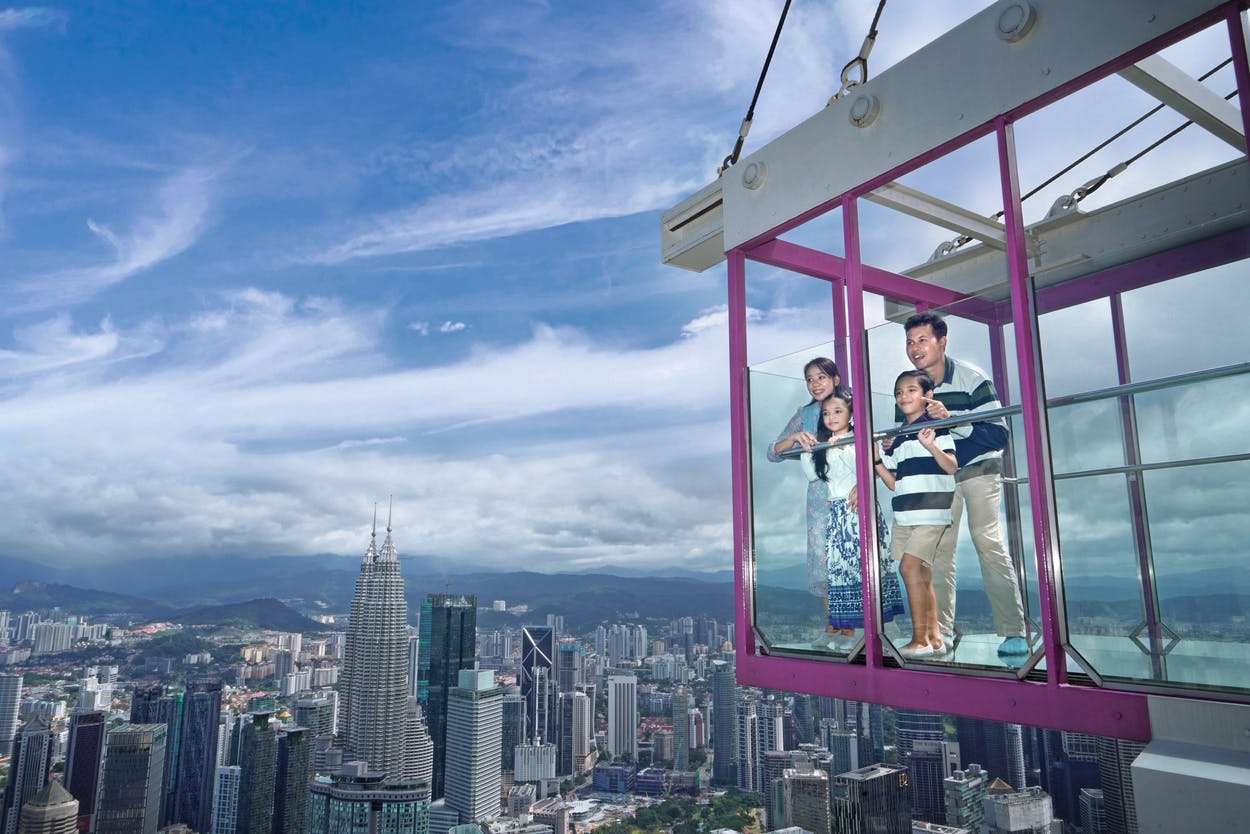 A family of four stands in a glass observation box high above a city skyline on a clear day.