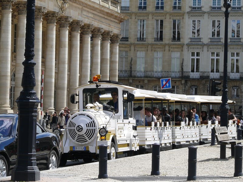 A white tourist train with passengers passes by historic buildings and columns in a European city.