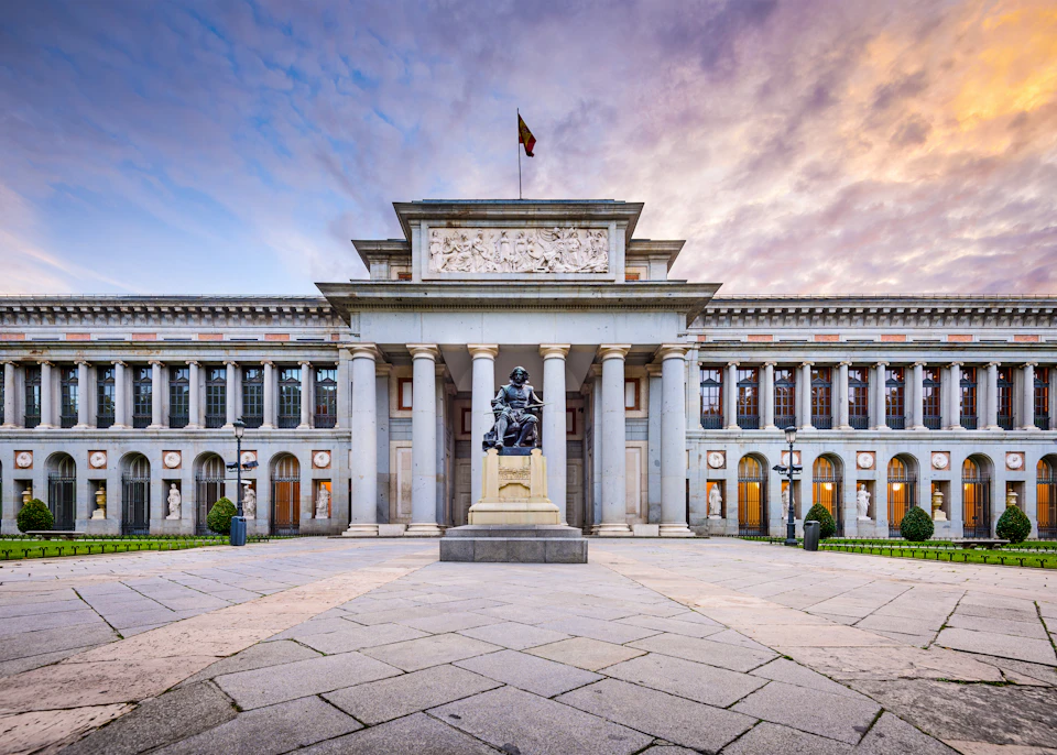 Grand building with columns, central statue, and detailed frieze under a flag, captured at sunset with a partly cloudy sky.