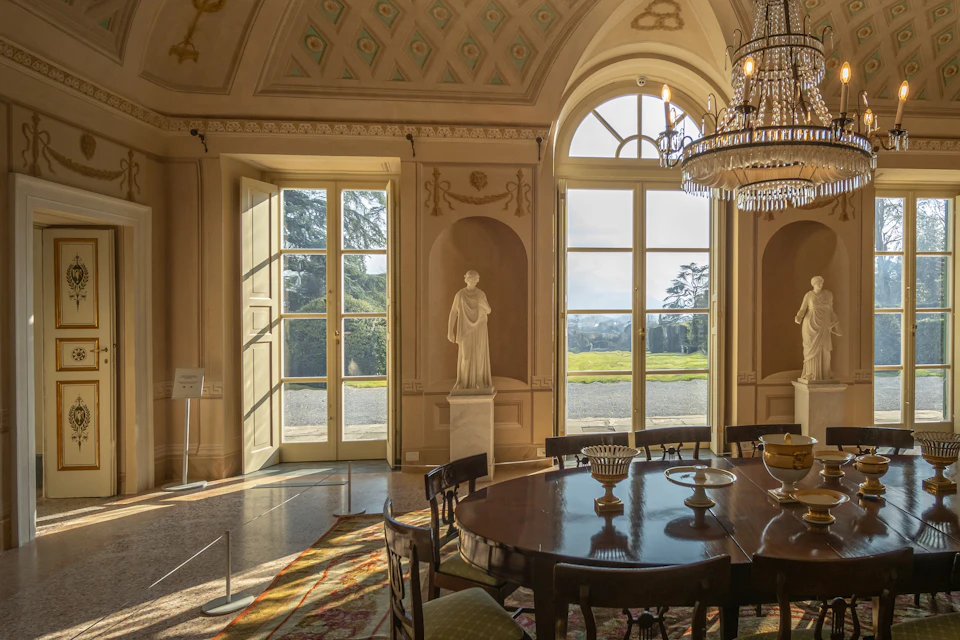 Elegant room with tall windows, a chandelier, a round dining table, and a statue on a pedestal near the window.