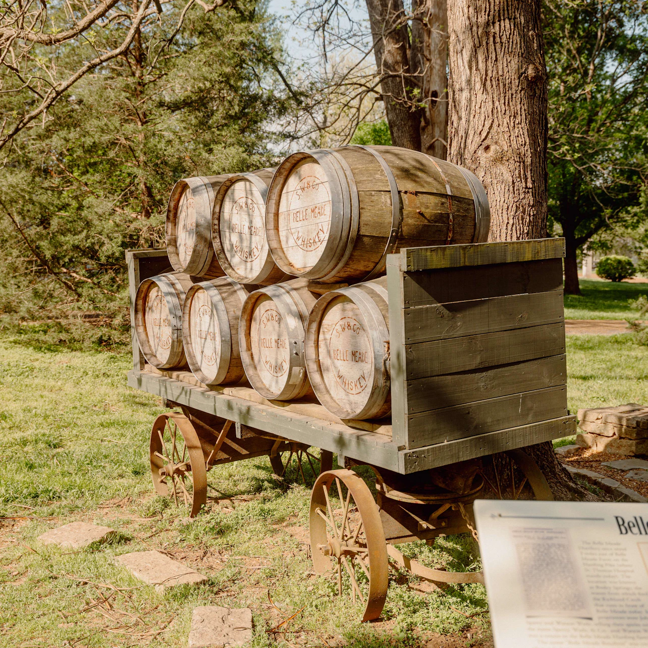 Wooden cart with six barrels in a grassy area, surrounded by trees. An information plaque is in the foreground.