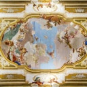 Baroque ceiling fresco featuring figures and clouds, surrounded by intricate gold and white architectural details.
