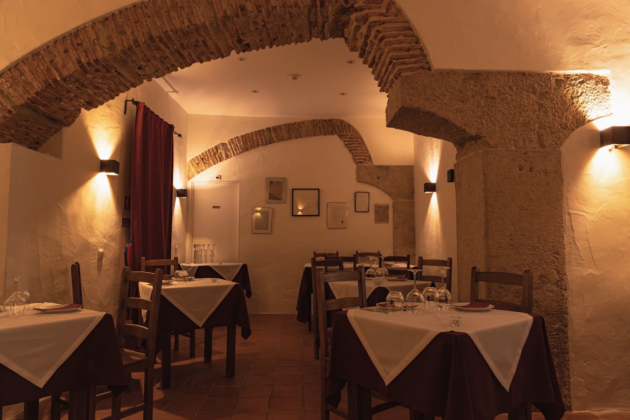 木製のテーブルと椅子、白いテーブルクロス、アーチ、額装された写真、テラコッタタイルの床がある薄暗いレストランのインテリア。