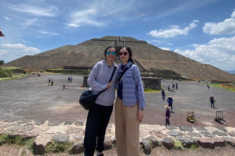 Two people pose in front of a large pyramid under a partly cloudy sky, with several other people walking around in the background.