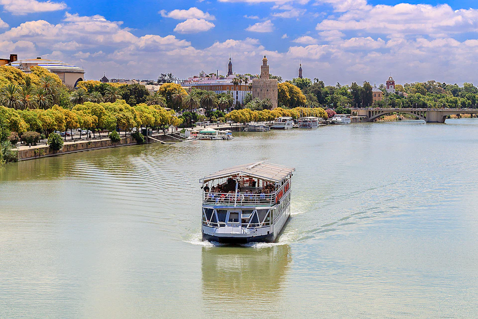 A boat sails on a river with lush greenery and historic buildings in the background, under a partly cloudy sky.