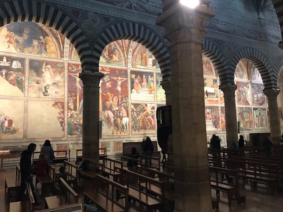 Wnętrze kościoła z misternymi łukami, freskami religijnymi i drewnianymi ławkami, z kilkoma osobami obserwującymi dzieło sztuki.