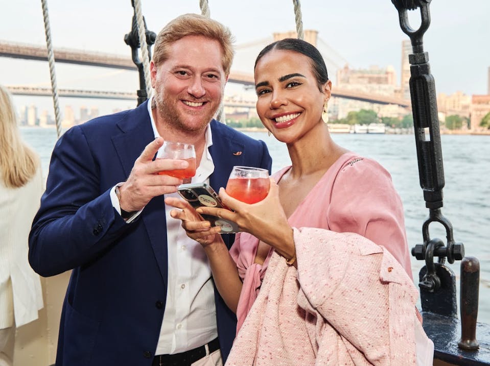 A man and a woman smiling while holding glasses of a pink drink. They are outdoors with a bridge and water in the background.