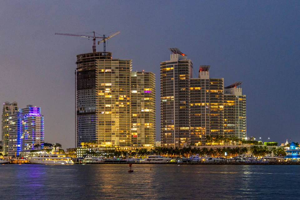 A waterfront cityscape at twilight showing lit high-rise buildings, a construction crane, and reflections on the water.